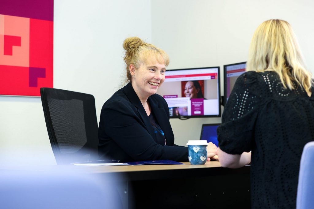 Smiling lady with red hair sits at desk and chats with woman with blonde hair